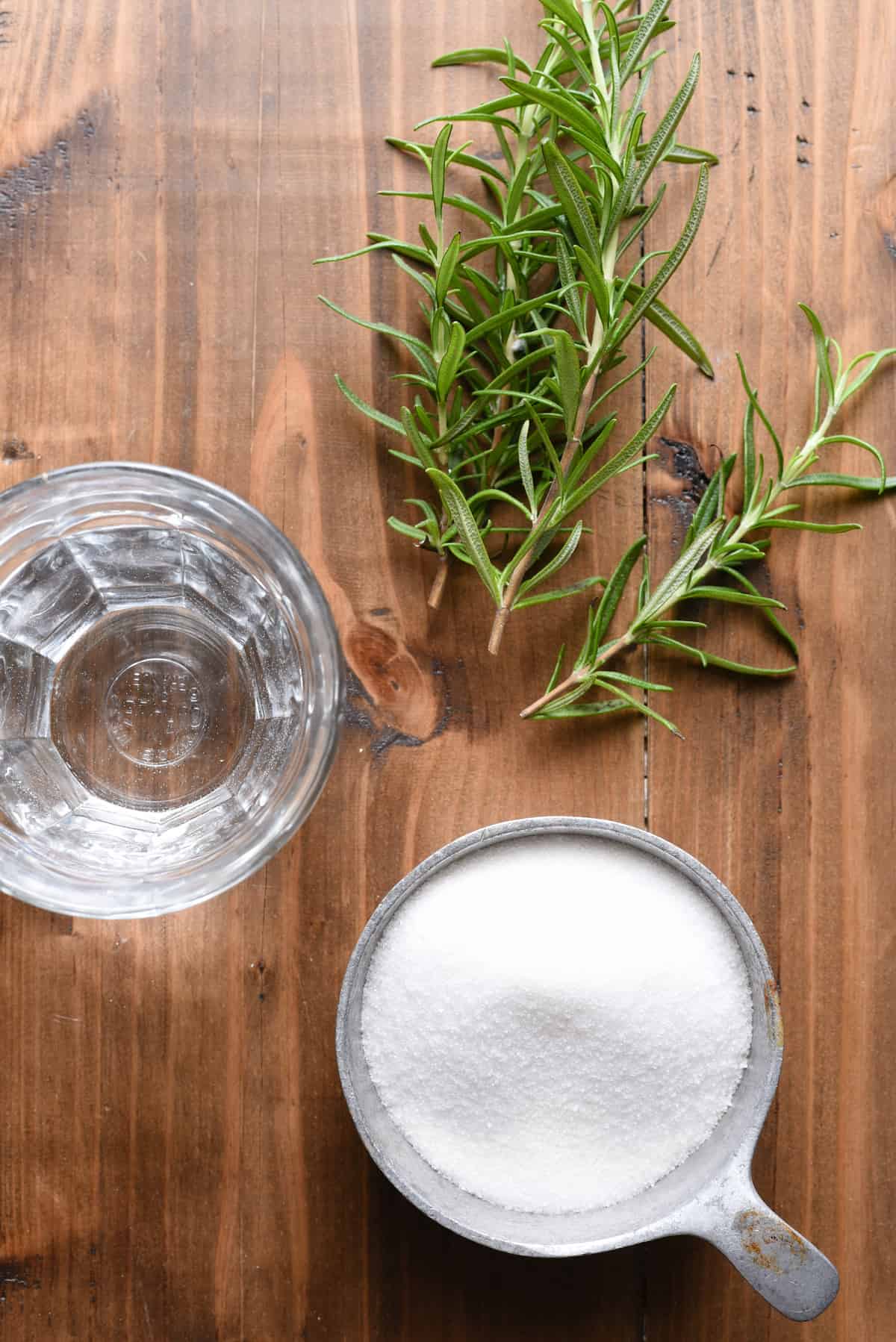 This homemade Rosemary Simple Syrup will make you feel like a fancy bartender, right in your own kitchen. Whip up a batch of this, and use it for whisky, gin, vodka or mocktails. | foxeslovelemons.com Overhead photo of cup of sugar, glass of water and rosemary sprigs on wooden table.