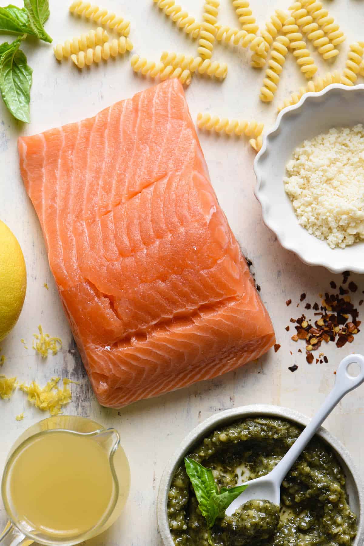 This Salmon Pesto Pasta is a restaurant quality dish that you can make at home. So flavorful, and easy enough to make on a weeknight! | foxeslovelemons.com Overhead photo of ingredients for salmon pesto pasta, including a salmon fillet, dried pasta, basil leaves, Parmesan cheese, chicken broth, basil pesto, lemon zest, and red pepper flakes.