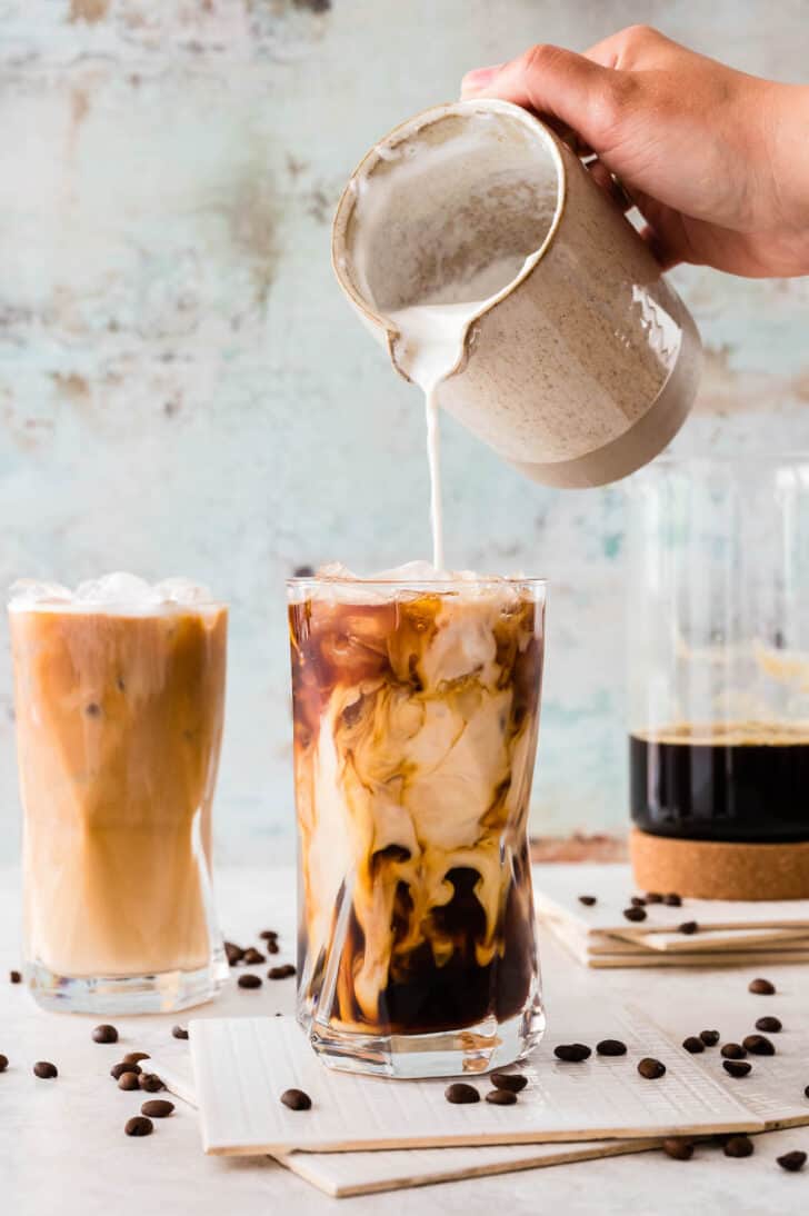 A large glass of iced coffee with cream from a pitcher being poured into it. The scene is garnished with napkins, a pitcher of coffee, and coffee beans.