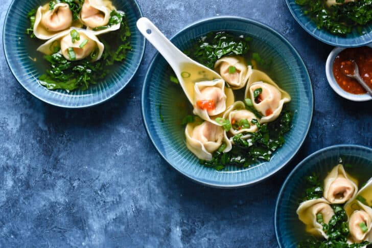 4 blue bowls filled with a wonton soup recipe on a blue background. A small bowl of hot sauce is nearby.