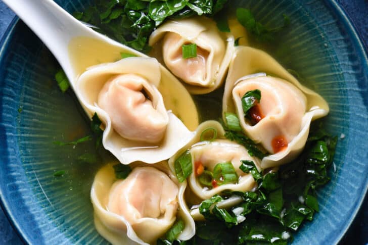 A broth with Chinese dumplings in a blue bowl with a white spoon scooping up one of five dumplings.