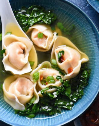 Shrimp wonton soup with kale in a blue bowl with a white soup spoon scooping up one of five wontons.