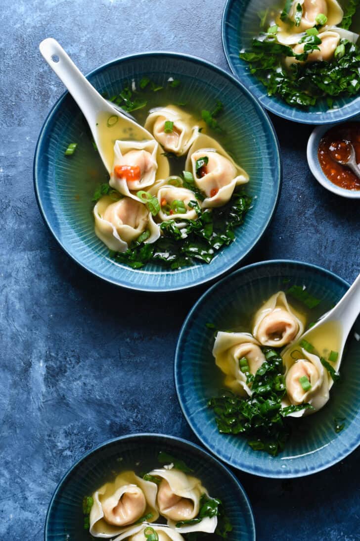 4 blue bowls are filled with homemade wonton soup. White soup spoons are scooping up wontons in 2 of the bowls.