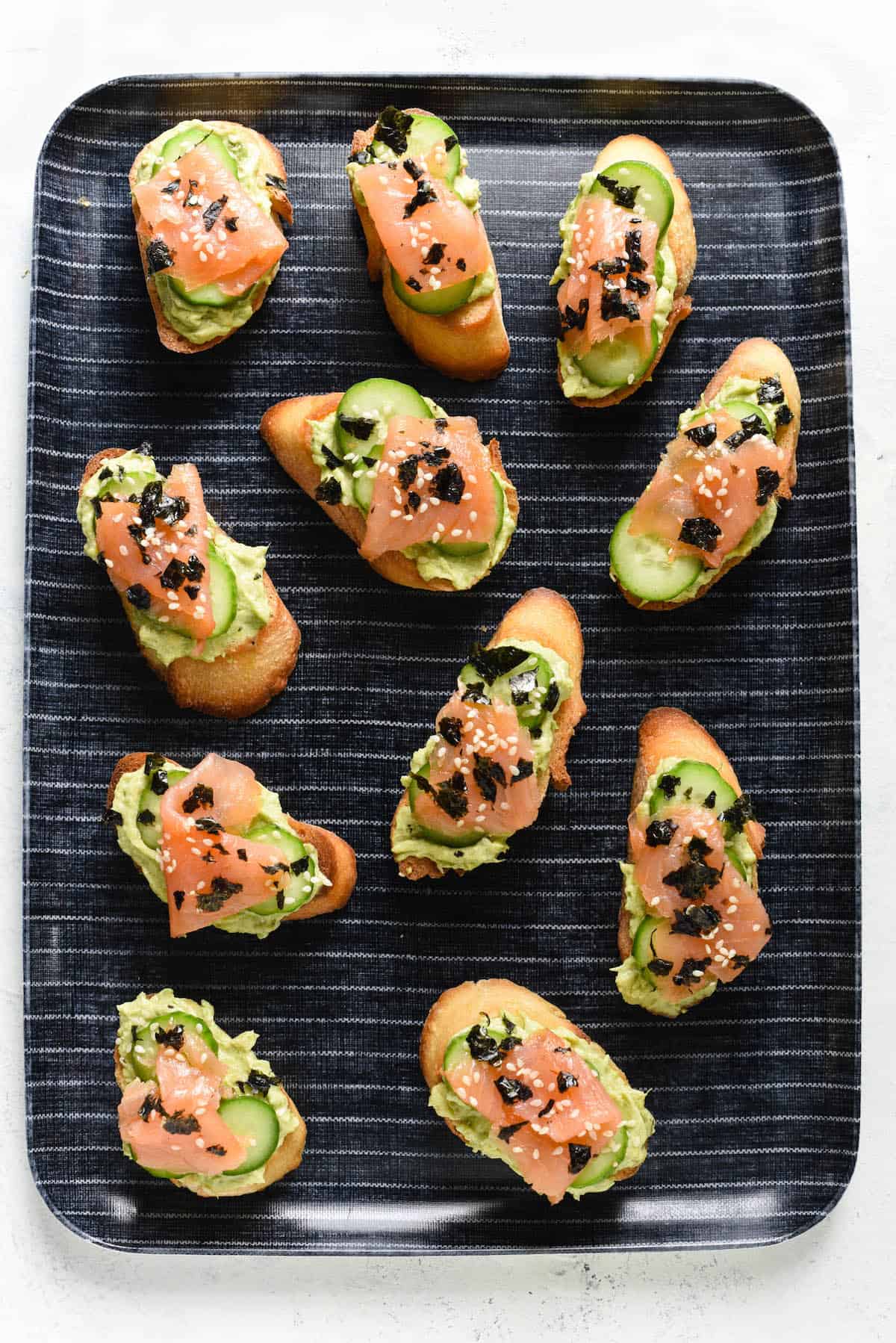 It's Smoked Salmon Crostini, with a twist. Avocado, cream cheese, cucumber and sesame seeds make this crostini taste just like a Philadelphia sushi roll! | foxeslovelemons.com Avocado, salmon and cucumber crostini arranged on blue and white striped tray.