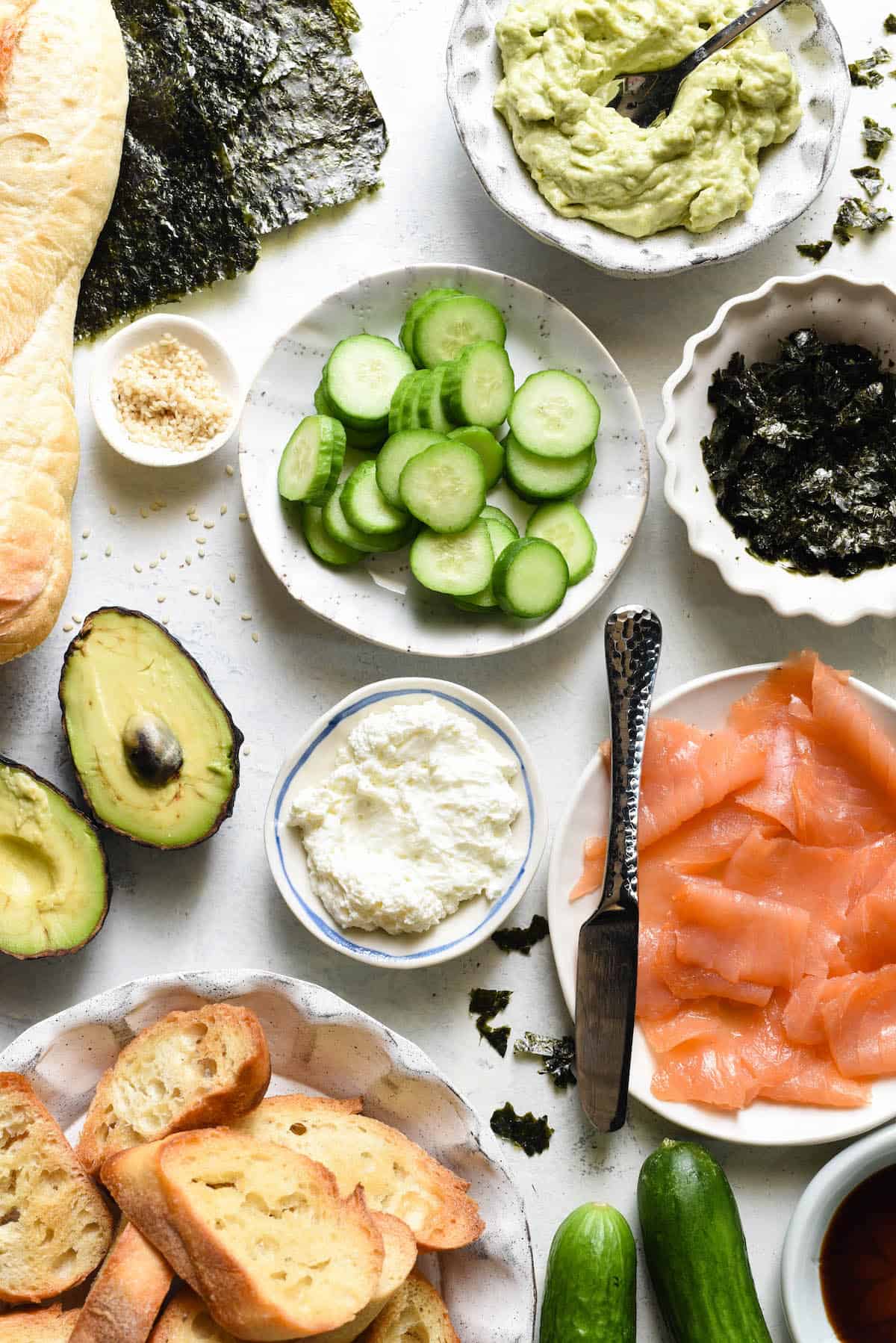 It's Smoked Salmon Crostini, with a twist. Avocado, cream cheese, cucumber and sesame seeds make this crostini taste just like a Philadelphia sushi roll! | foxeslovelemons.com Ingredients for salmon bruschetta arranged in small bowls on white tabletop. Avocado spread, sliced cucumber, seaweed, smoked salmon, cream cheese, sesame seeds, sliced baguette.
