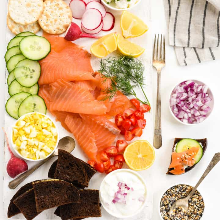 White table and cutting board topped with smoked salmon and garnishes like cucumber, capers, vegetables, cream cheese, bread and crackers.