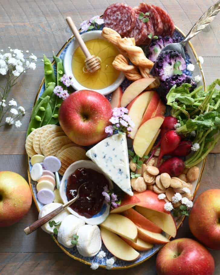 Oval platter filled with cheeses, fruit, honey, jam, nuts, meats and flowers.