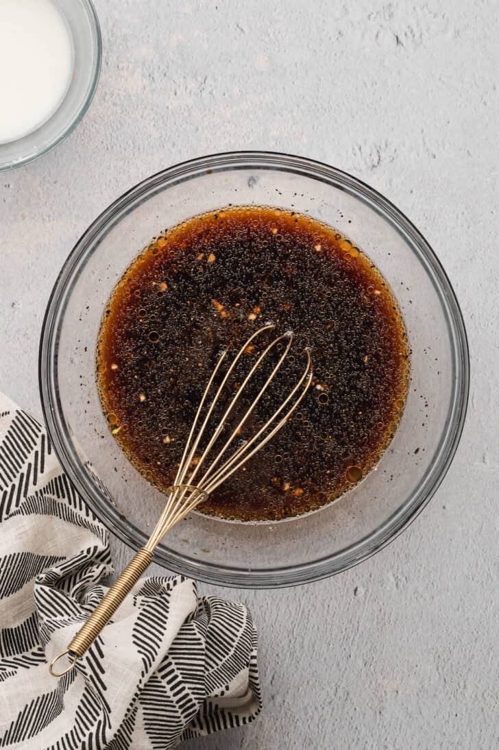A glass bowl with a metal whisk stirring together a brown liquid.