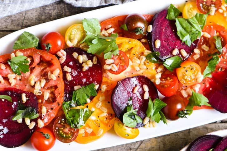 A beet and tomato salad on a white platter garnished with herbs and barley.