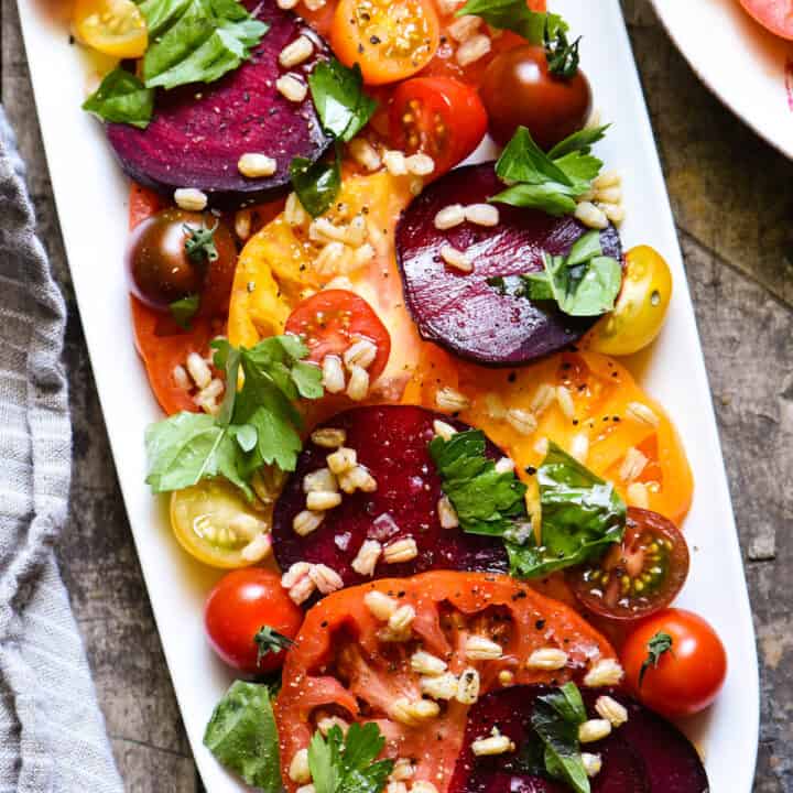 White oblong platter filled with sliced tomatoes, beets, herbs and barley.