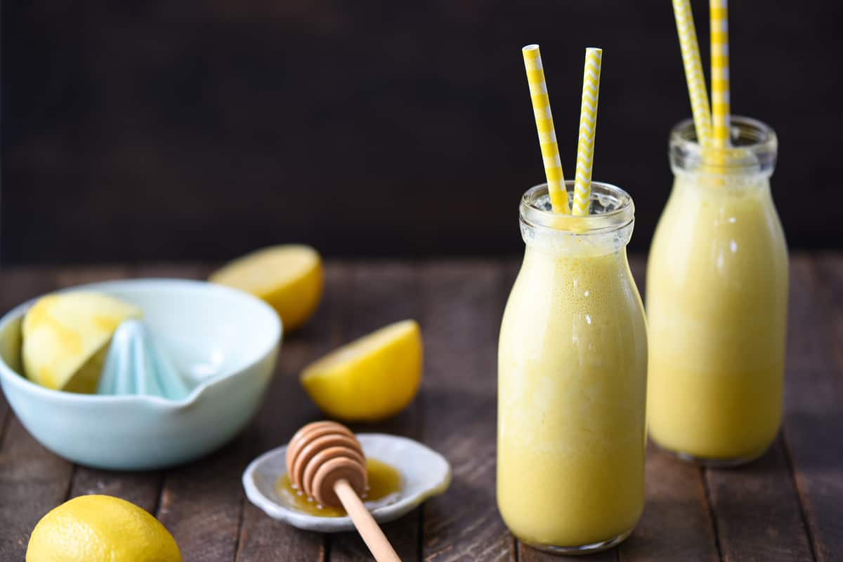 This Lemon Smoothie is like sunshine in a bottle! Yogurt, lemon, honey, ginger and turmeric will wake you up on a sleepy morning. | foxeslovelemons.com Two small glass bottles filled with lemon ginger smoothie, with yellow straws sticking out of the bottles. A citrus juicer and a honey dish are in the background.