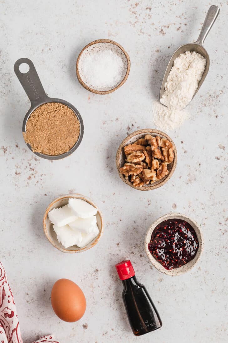 Ingredients on a light surface, including flour, salt, brown sugar, walnuts, shortening, jam, an egg and vanilla extract.