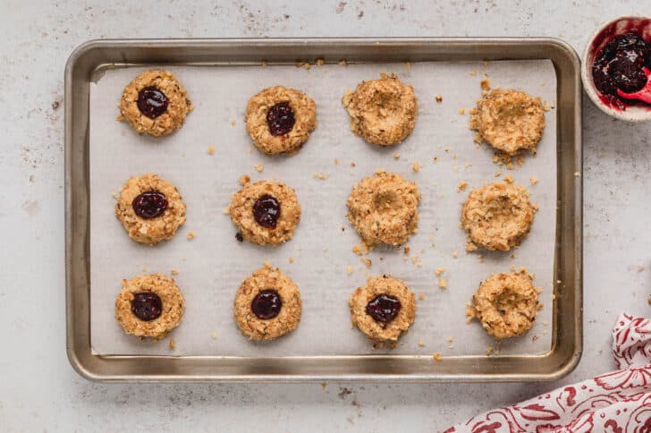 Jam thumbprint cookies on a rimmed baking pan, in process of being filled with jam.