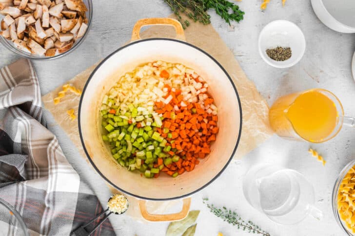 A white Dutch oven with diced onions, carrots and celery sauteeing in it.