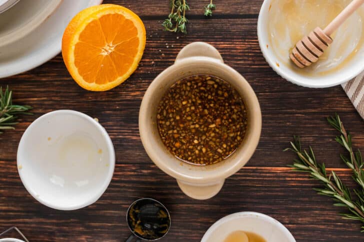 A small tan bowl filled with a mixture of soy sauce and garlic, with a fresh orange half and herbs nearby.