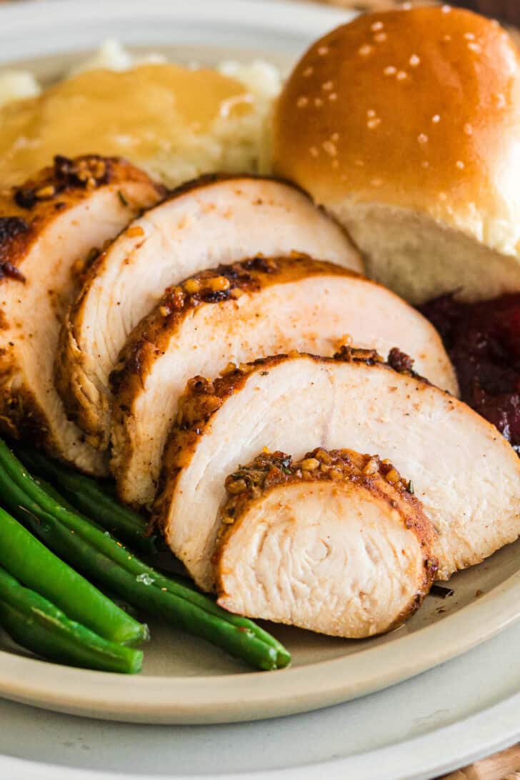 A turkey tenderloin recipe, sliced, on a plate with green beans, mashed potatoes and gravy, a roll and cranberry sauce.