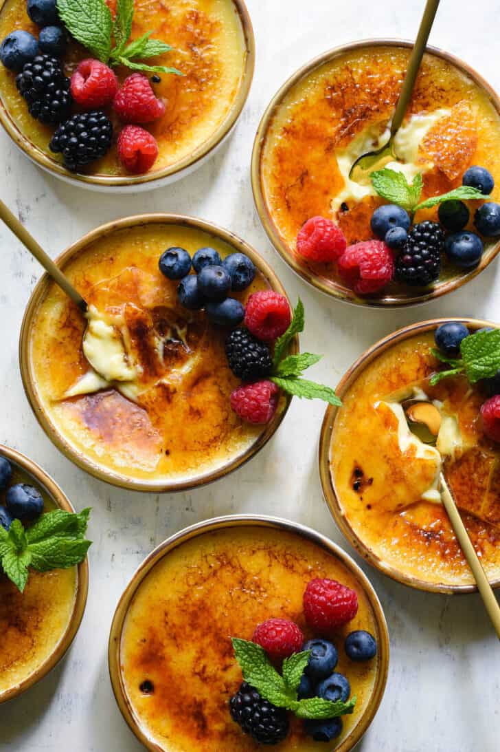 Six dishes of vanilla bean creme brulee garnished with berries and mint sprigs, on a light surface, with gold spoons digging into three of the dishes.