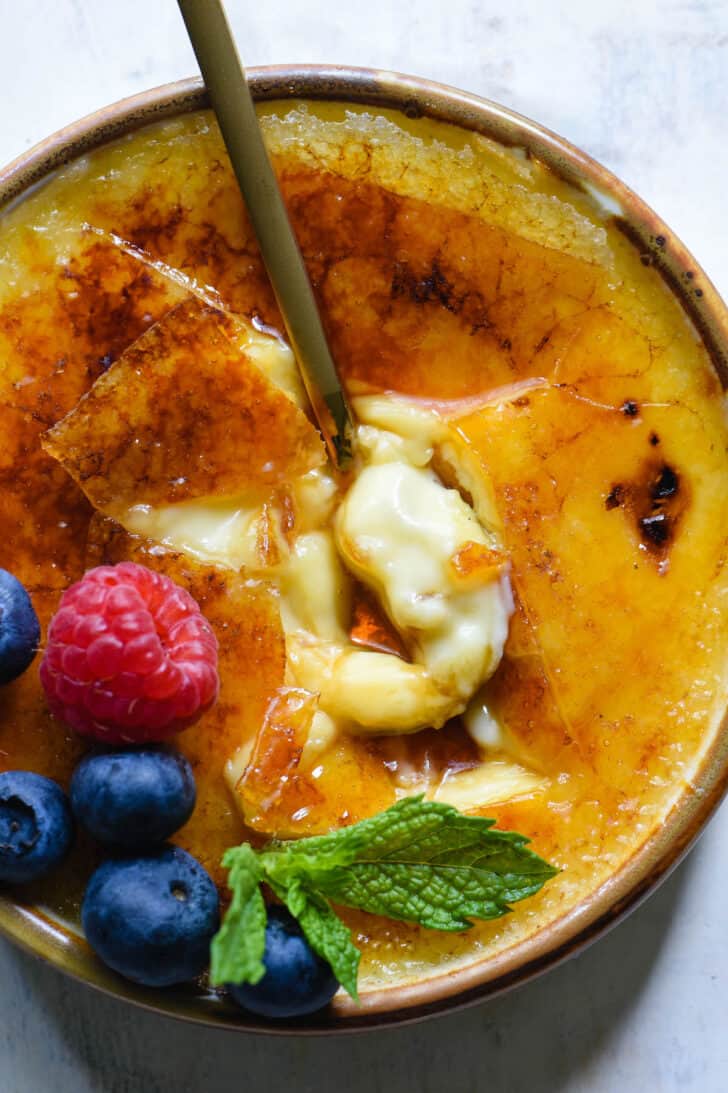 Closeup on a gold-rimmed ramekin of vanilla creme brulee garnished with blueberries, raspberries, a blackberry and a mint sprig, with a gold spoon breaking through the torched sugar.