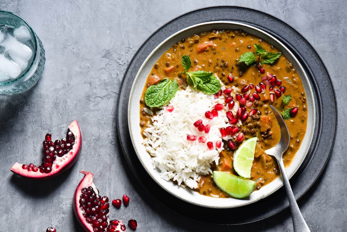 This Vegan Lentil Curry is a healthful one-pot meal packed with layers of flavor, using ingredients easy to find at your grocery store. Perfect for a meatless Monday dinner, or any time your meal planning could use a reset button. | foxeslovelemons.com Bowl of vegan curry and white rice garnished with lime wedges, pomegranate seeds and mint leaves.