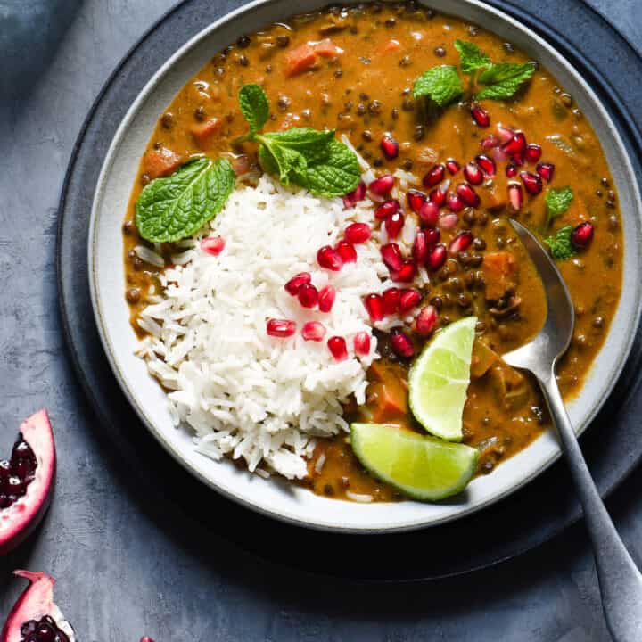 Bowl of vegan lentil curry and white rice garnished with lime wedges, pomegranate seeds and mint leaves.