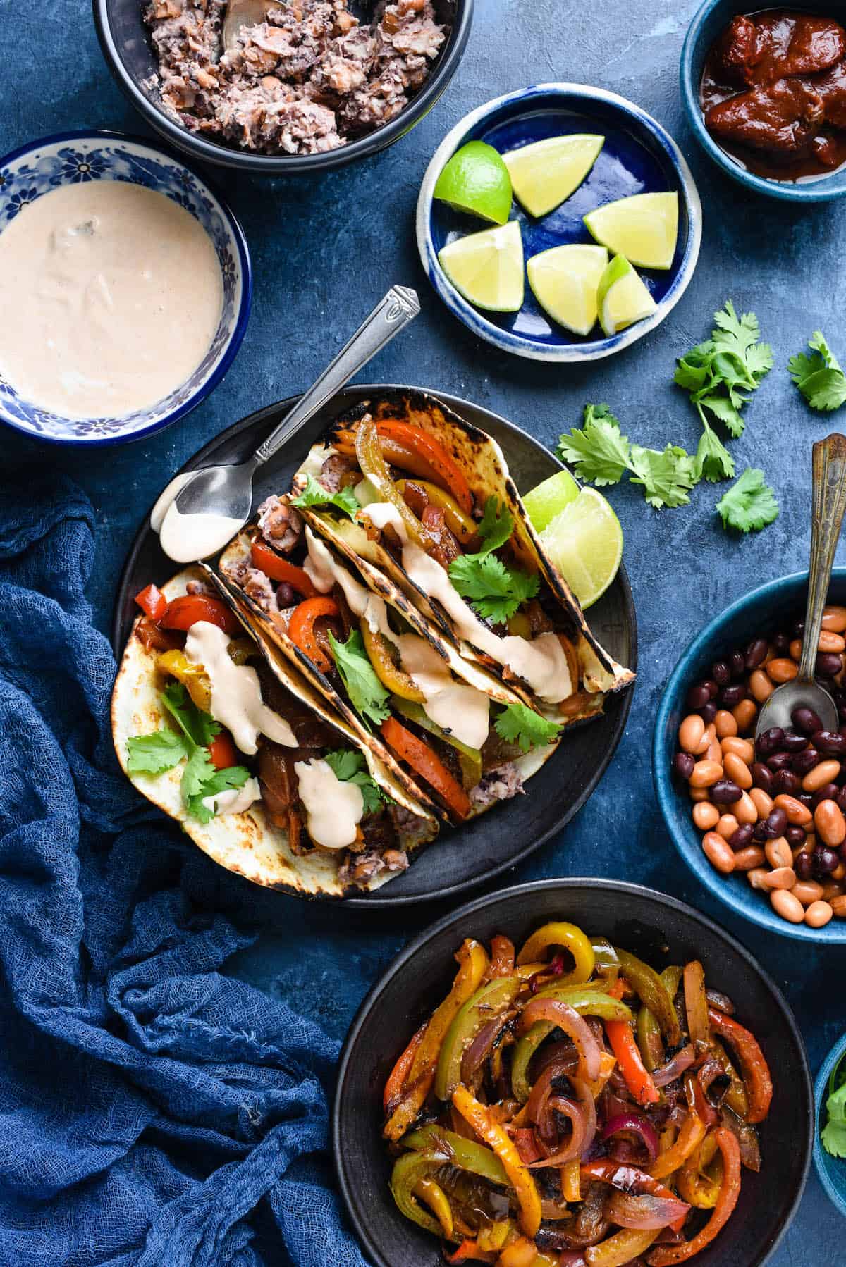 These Vegetarian Fajitas with Creamy Chipotle Sauce are a meatless but protein-packed meal with a delicious restaurant-style sauce! | foxeslovelemons.com Overhead photo of plate of vegetarian fajijtas, along with bowls of creamy chipotle sauce, mashed beans, lime wedges, chipotles in adobo sauce, whole black beans and pinto beans, and cooked fajita vegetables.