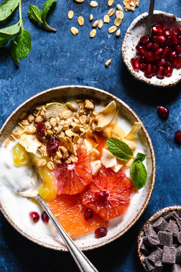 A textured bowl filled with yoghurt and topped with sliced oranges, granola, lemon curd, mint and pomegranate seeds.